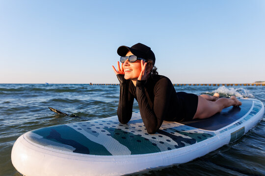 Smiling woman enjoys surfing on Baltic Sea waves, laughing joyfully lying on board with splashing water around. Bright sunlight and her laughter reflect pure joy, freedom and vibrant summer adventure - Powered by Adobe