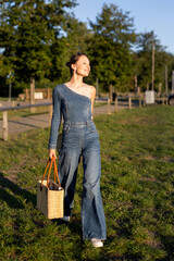 Young adult beautiful woman wear stylish jeans one-shoulder denim jumpsuit have fun walks outdoors warm sunny day holding straw bag hat hand. Female person carefree smiles enjoying countryside nature