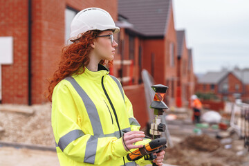 Portrait of confident female land surveyor with long hair in bright safety gear using surveying instrument to perform land survey on building site