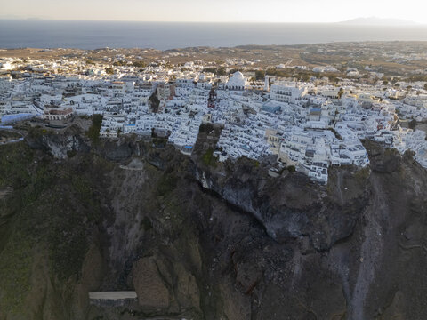 Aerial view of whitewashed buildings cascading down rugged cliffs, meeting the azure sea under a golden sky, Fira, Santorini, Greece.