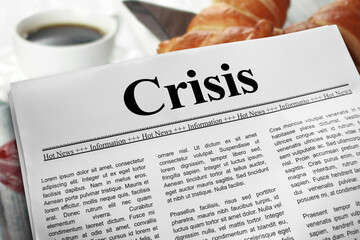 Crisis in newspaper layout on a breakfast table with a blurry cup of coffee and croissants in the background.