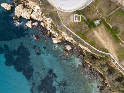 Aerial view of turquoise waters meeting the rugged coastline near a white structure, contrasting with the earthy tones of the landscape, Paros, Paros, Greece.