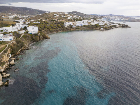 Aerial view of the shoreline where turquoise waters meet the rocky coast near whitewashed buildings, a serene blend of natural beauty and Cycladic architecture, Paros, Paros, Greece.