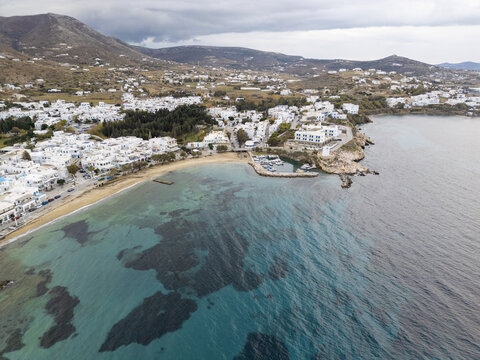 Aerial view of the coastline's embrace, where white buildings meet the turquoise sea, and dramatic cliffs rise against the skyline, Paros, Paros, Greece.
