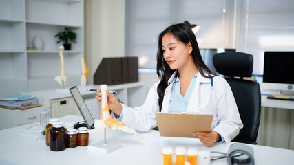 Asian Woman Doctor at Desk with Spinal Model tablet in Clinic for Healthcare Treatment Analysis and Patient Education Explaining Diagnosis