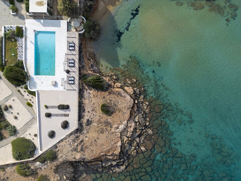 Aerial view of turquoise waters meet rocky shores beside a pristine white villa with a shimmering pool, a luxurious contrast of nature and architecture, Paros, Paros, Greece.