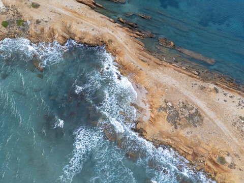 Aerial view of the rugged coastline where the turquoise sea crashes against the golden cliffs, creating a stunning contrast of colors and textures, Paros, Paros, Greece.