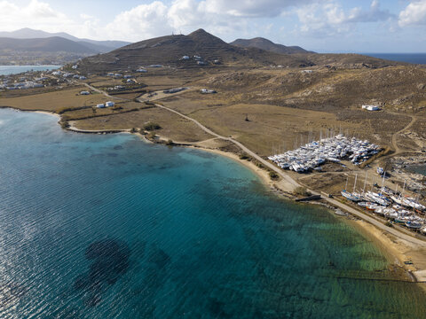 Aerial view of turquoise waters meet the arid landscape where a marina of white boats rests near brown hills, Paros, Paros, Greece.