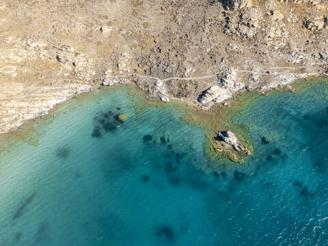 Aerial view of turquoise waters meeting the rugged coastline, a dance of vibrant blues and earthy browns, revealing the serene beauty of the coast, Paros, Paros, Greece.