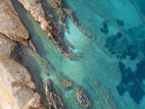 Aerial view of jagged, sun-kissed rocks meet the turquoise embrace of the Aegean Sea, creating a mesmerizing contrast of textures and tones, Paros, Paros, Greece.
