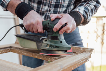 Craftsman. Adult carpenter using an electric sander to smooth an old wooden window. Construction industry, carpentry, housework do it yourself. Restoration.