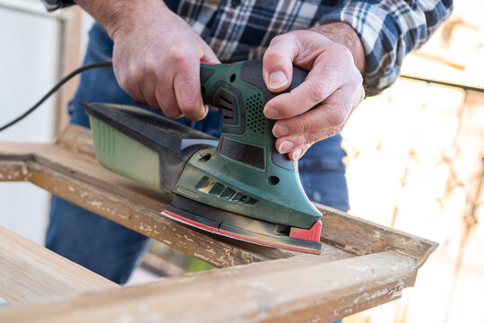 Craftsman. Adult carpenter using an electric sander to smooth an old wooden window. Construction industry, carpentry, housework do it yourself. Restoration.