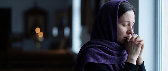 Devout woman wearing purple headscarf praying with clasped hands in church. Christian concept of Lent, faith and repentance. Spiritual portrait near window with icons in background.