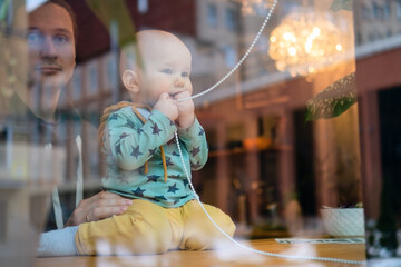 Young father with toddler in cafe holding child, visible through window. Family values, parenting love, bonding and care in public place with warm atmosphere and happiness.