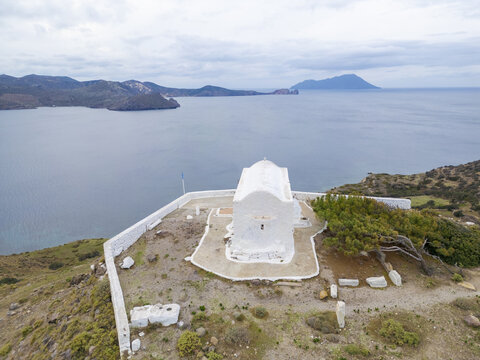 Aerial view of the stark white Prophet Elias Holy Chapel perched atop a hill, contrasting against the deep blue sea and cloudy sky, Prophet Elias Holy Chapel, Milos, Greece.