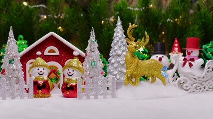A fairytale Christmas village with a house surrounded by snowmen and Christmas trees behind a fence and a deer with snowmen nearby against the backdrop of large Christmas tree branches