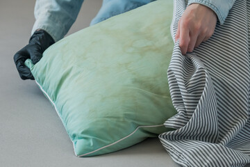 A person wearing a black glove touching a green pillow with dirty yellow stains sweat or saliva in a living room. Conscious home care and sustainable living concept. top view