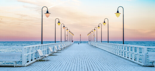 Wide view of an empty wooden pier stretching into the sea during colorful sunset. Street lamps glow...