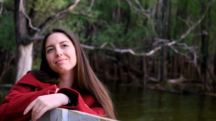 Portrait photo of beautiful young smiling woman in burgundy coat sitting on bench outdoors in park.