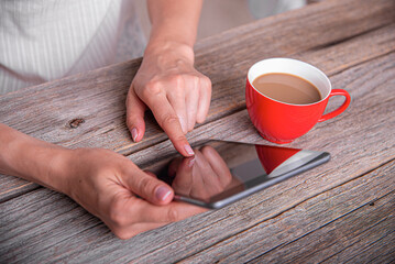 A woman uses a digital tablet on a rustic wooden table next to a red cup of coffee.