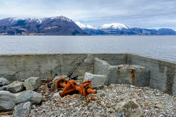 Rusty equipment sits on rocky shore against a backdrop of mountains and cloudy sky