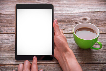 A woman's hands holding a digital tablet with a blank white screen next to a cup of coffee on a rustic wooden table.