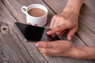 A woman's hands hold and interact with a black smartphone on a rustic wooden table next to a cup of coffee.