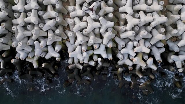 Conceptual aerial video of a lone person lying among massive concrete tetrapod breakwater blocks. Human vulnerability and isolation against monumental coastal infrastructure and industrial geometry