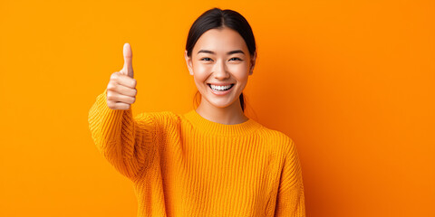 Happy young Asian woman in orange sweater smiling and showing a thumbs up gesture against a bright orange background.