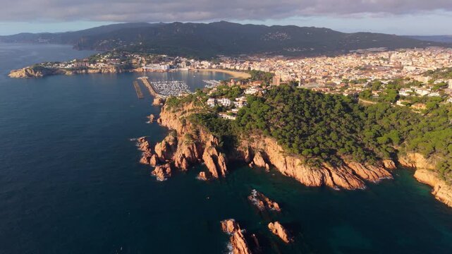 Stunning aerial perspective flying over the rugged cliffs toward the coastal town of sant feliu de guixols in costa brava, spain, capturing the marina and beach at sunrise