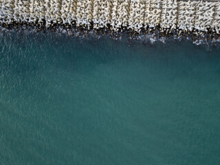 Top-down aerial view of massive concrete wave breakers meeting the sea, showing contrast between rigid human-made infrastructure and powerful natural water, forming an abstract, graphic coastal