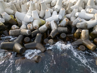 Top-down aerial view of massive concrete wave breakers meeting the sea, showing contrast between rigid human-made infrastructure and powerful natural water, forming an abstract, graphic coastal