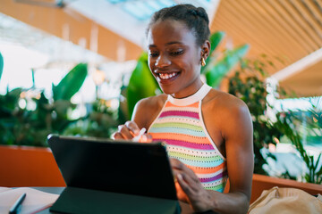 Young woman smiling at digital tablet with stylus in hand, enjoying tech-enabled creative moment in lush indoor workspace with daylight and vibrant greenery.