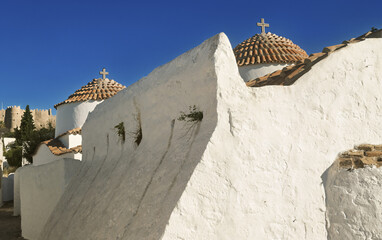 Street scene in Patmos, Greece, showing a church and a narrow passage lined with whitewashed stone buildings and flat roofs, typical of Cycladic architecture. 