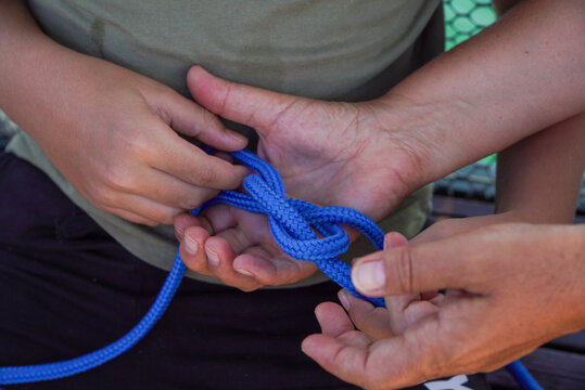 A close-up of hands tying a knot with bright blue rope. The image symbolizes teamwork, cooperation, connectedness, trust, and practical skills.