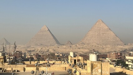 Pyramids rise in Giza with buildings nearby. The scene shows a mix of modern structures and ancient wonders under a dusty sky. People visit the site as history meets current life