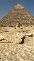 Visitors gather around the base of a large pyramid in Giza, Egypt. The ground is dry with rocks scattered around. The pyramid rises sharply into the blue sky above as tourists look up in awe