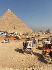 Groups of people visit the Giza pyramids in Egypt while horse-drawn carriages are parked nearby. The sun shines brightly as tourists walk and enjoy the historical site