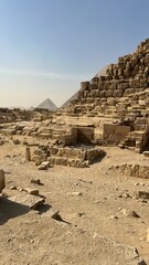Visitors walk among large stone blocks and ruins at the Giza Plateau. The sun shines brightly overhead as the well-known pyramids stand in the background. Dusty ground is visible