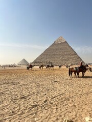Tourists are on horseback near the Great Pyramid in Giza. The scene shows several pyramids and a clear blue sky. The sandy landscape surrounds the riders and their horses