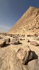 A pyramid stands tall in Egypt with a slope of stones. The ground is scattered with rocks, and the sky is blue above. Sunlight highlights the ancient stones