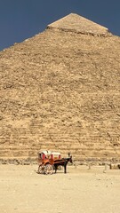 A horse cart stands still near the Great Pyramid in Giza. A person is in position by the cart as the sun shines down on the sandy ground. The pyramid looms large in the background