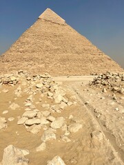 People walk near the Great Pyramid of Giza in Egypt. The pyramid stands tall with a clear blue sky above. Dusty ground surrounds the ancient structure with stones scattered around it