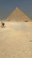 People ride horses in the desert near the pyramids of Giza. The scene shows dust rising as horses move along the sandy path under a bright sky with sunlight shining down