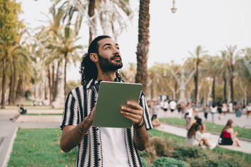 Confident young man holding tablet looks up in palm-lined park, showing curiosity, digital engagement and peaceful connection with modern tech outdoors. © BullRun