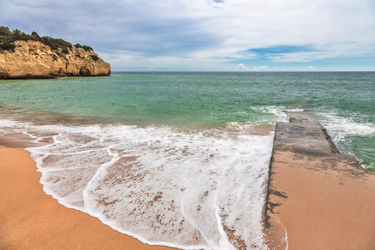 A serene coastal scene in the Algarve, Portugal, featuring a weathered concrete pier extending into the turquoise waters of the Atlantic Ocean.