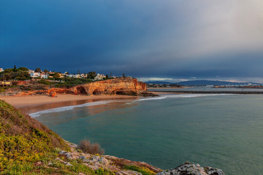 A dramatic and atmospheric view of Praia do Pintadinho in the Algarve, Portugal, captured during the golden hour under a moody, stormy sky.