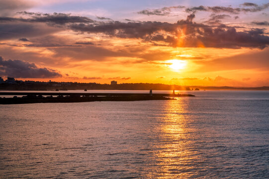 A stunning sunset over the harbor at Praia do Pintadinho, Algarve, Portugal, where the setting sun casts brilliant golden rays through the clouds, illuminating the water and silhouetting the breakwate