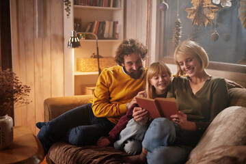 A family sits on a sofa in a comfortable living room, sharing a book while enjoying a cozy atmosphere filled with soft lighting and seasonal decorations.