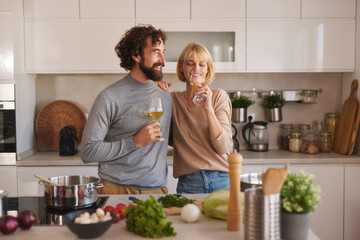 A couple stands in a stylish kitchen, smiling and holding glasses of wine as they prepare a meal. Fresh ingredients like vegetables and herbs surround them on the counter.